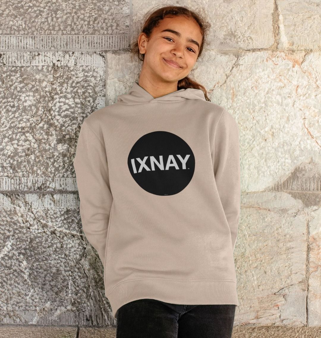 Girl smiling and leaning against old concrete block wall wearing kids oat light beige organic relaxed fit pullover hoodie with original black round IXNAY logo