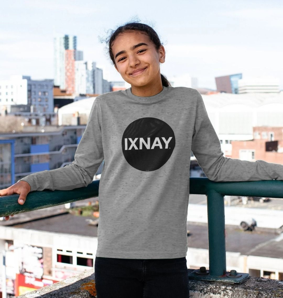 Girl smiling while standing on the top deck of a city parking garage and wearing a kids light gray organic long-sleeved t-shirt with original black round IXNAY logo

