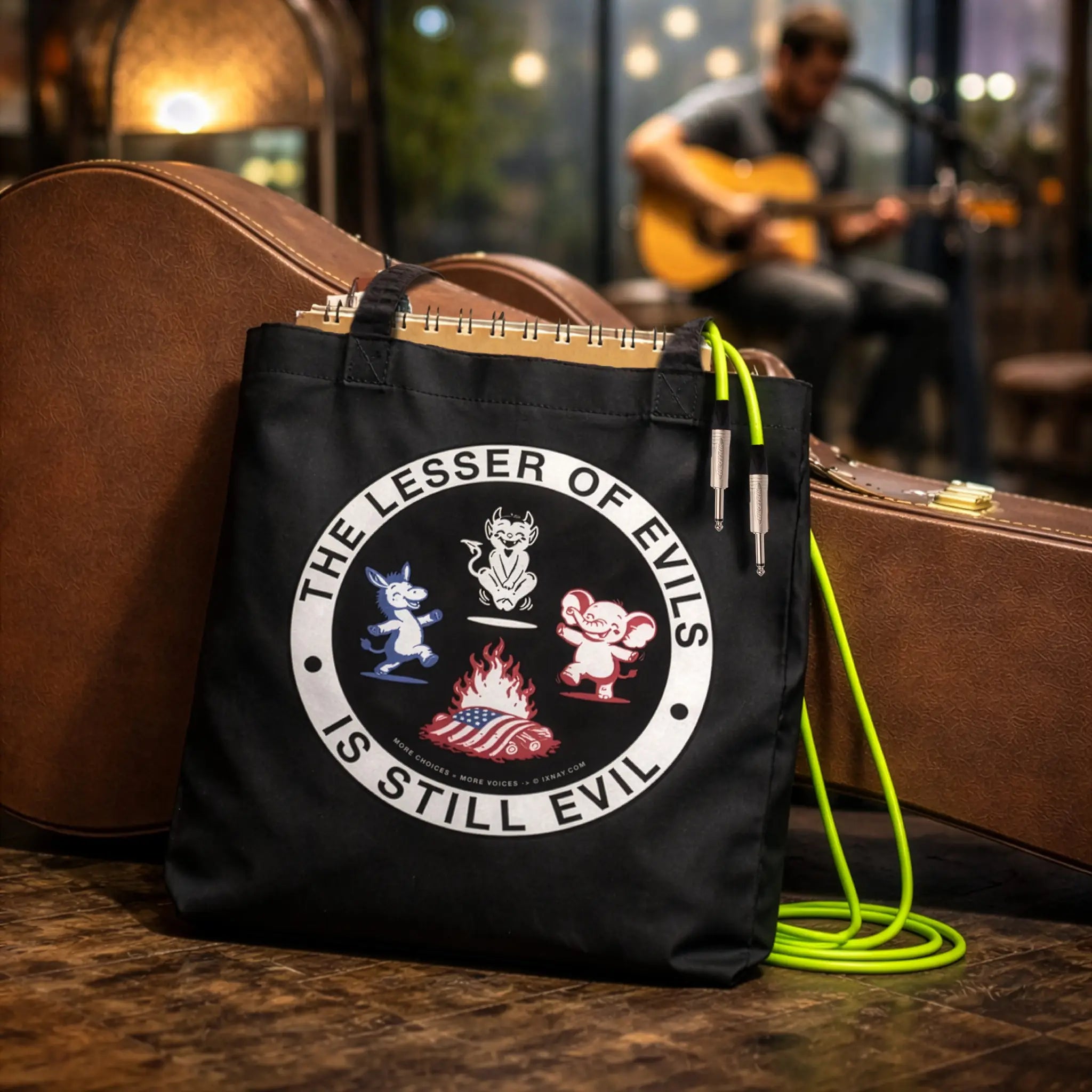 Black organic cotton tote bag with IXNAY's The Lesser of Evil is Still Evil graphic on front panel carrying music gear at open mic gig.