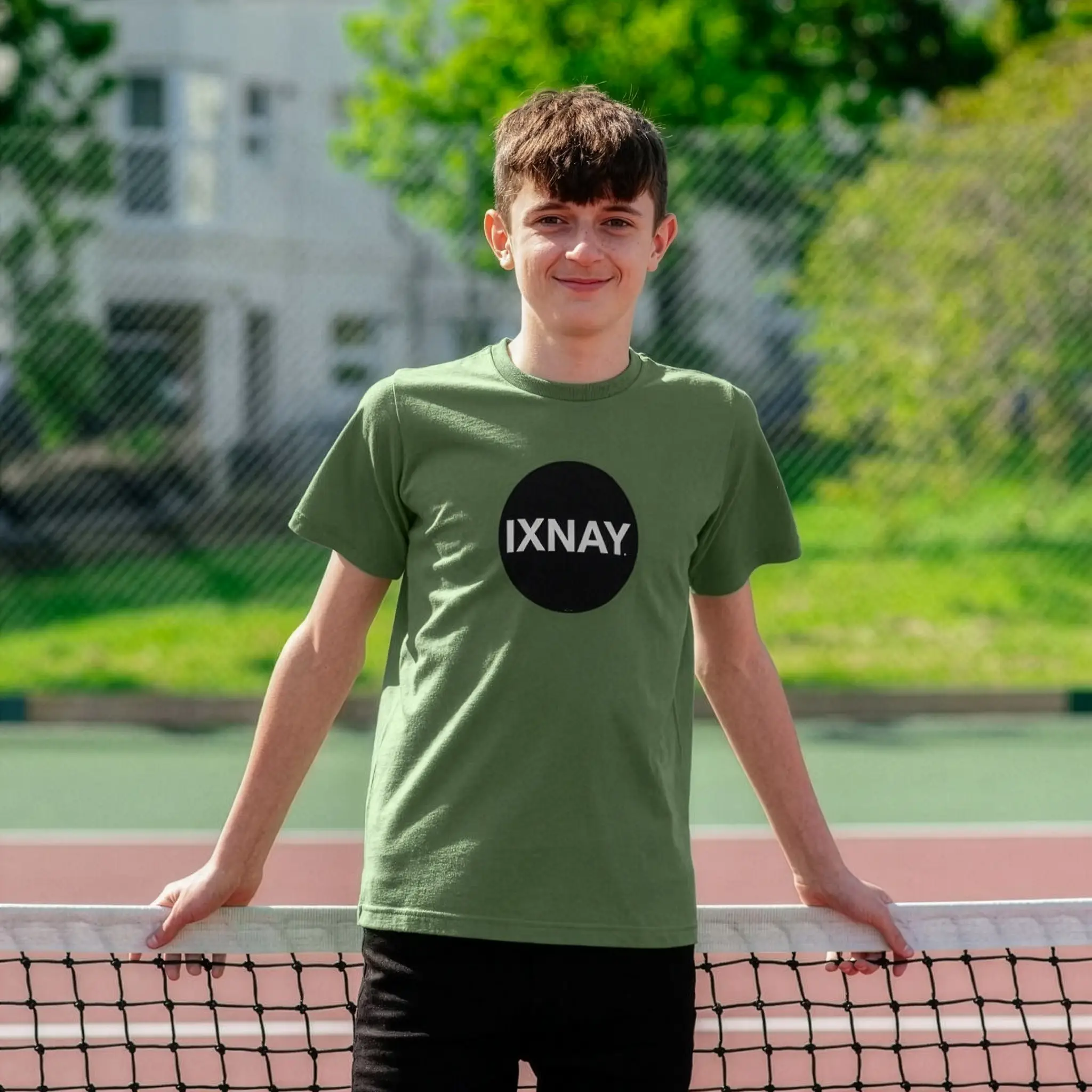 Young teen leaning against tennis net wearing IXNAY Kids 100% Organic Cotton T-Shirt Original Logo - Sage green.