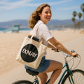 Woman riding a bike on a beach with an IXNAY Original Logo Canvas Tote Bag - 100% Organic Cotton.