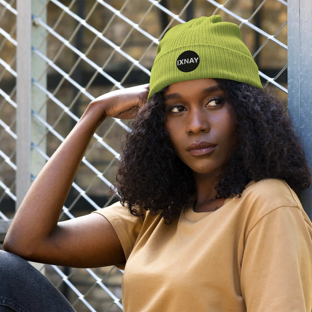 Woman wearing the IXNAY Original Logo 100% organic ribbed beanie in leaf green leaning against a chain link fence