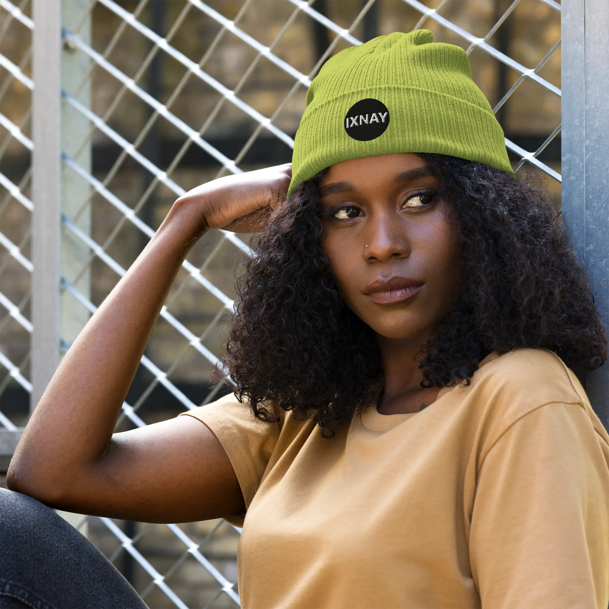 Woman wearing the IXNAY Original Logo 100% organic ribbed beanie in leaf green leaning against a chain link fence