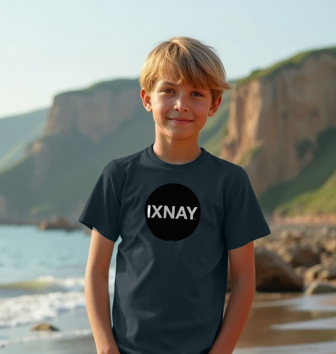 Smiling boy on ocean beach with cliffs in the background wearing a dark denim blue t-shirt with the original IXNAY black circular logo printed on the front