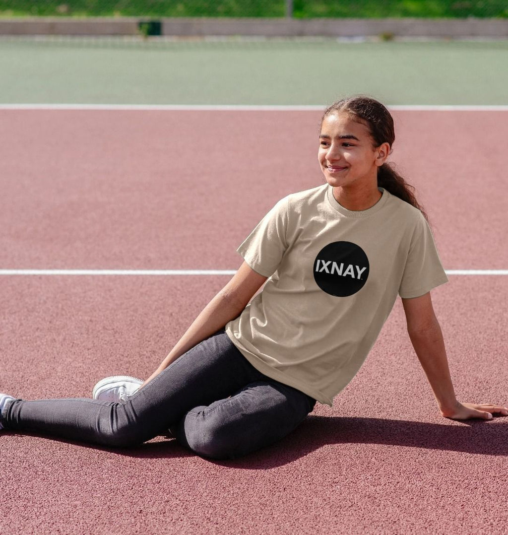 Smiling mixed race girl sitting and relaxing on a tennis court wearing an oat light beige t-shirt with the original IXNAY black circular logo printed on the front
