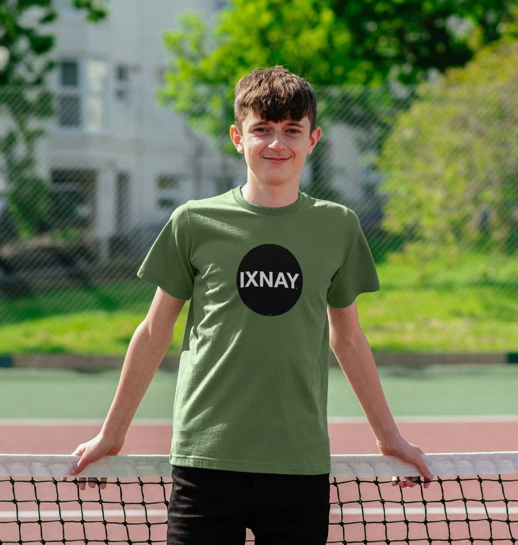 Smiling pre-teen kid leaning against the net on a tennis court wearing an sage greene t-shirt with the original IXNAY black circular logo printed on the front