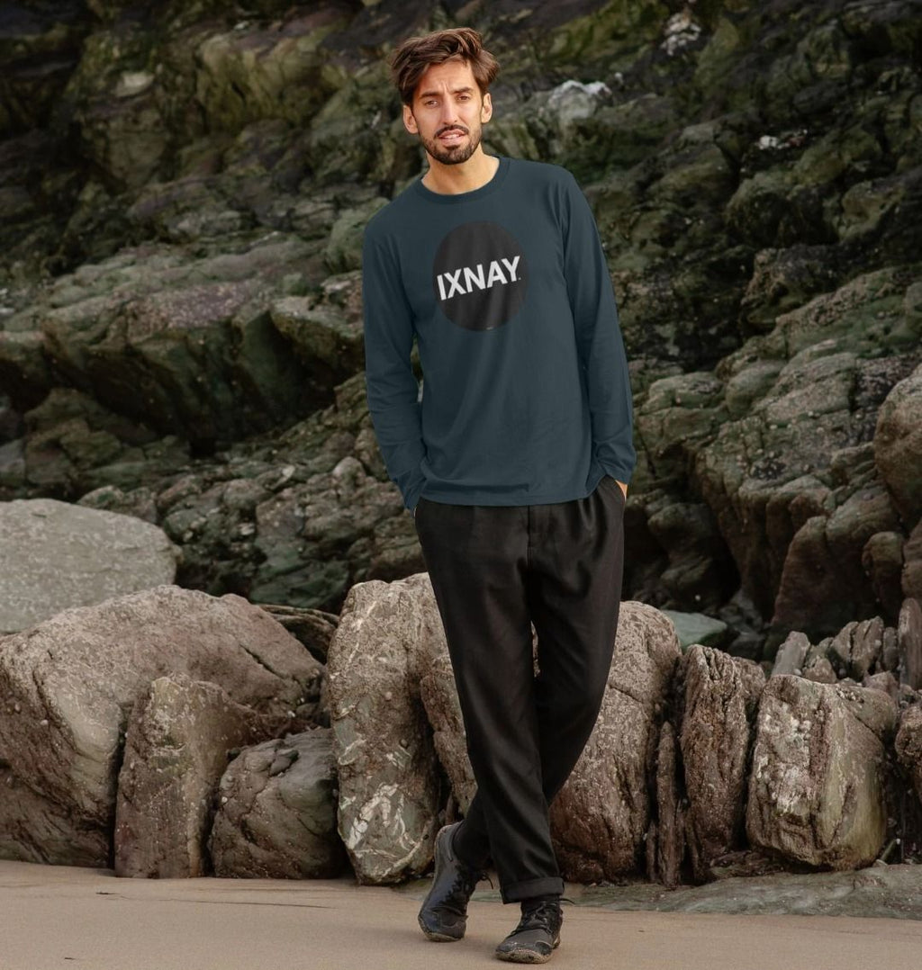 Man standing in front of large rock formation at the beach wearing Dark Denim Blue Printed Men's Long-sleeved T-Shirt with Original Black Round IXNAY Logo from the artist and musician IXNAY
