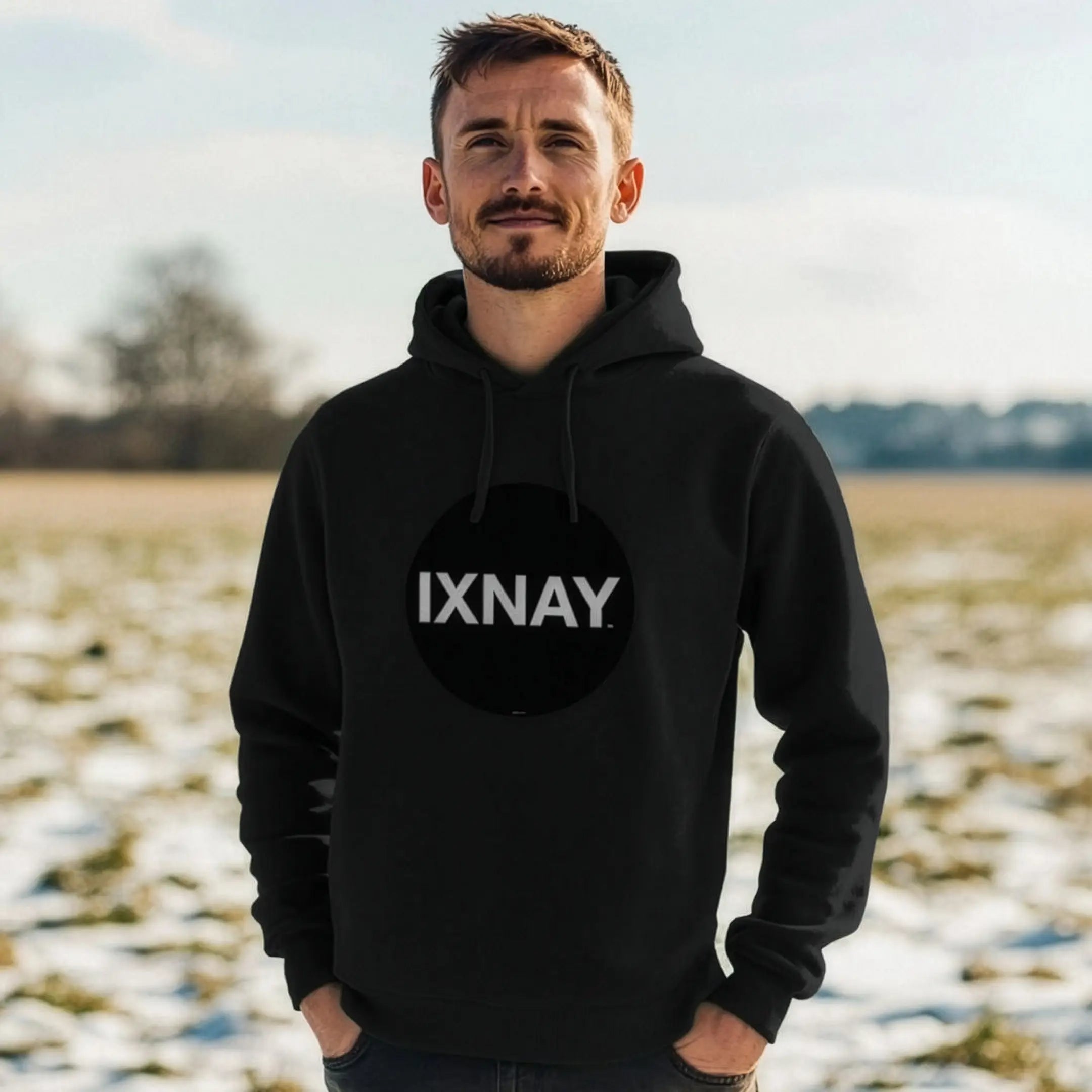 Man in partially snowy field wearing mens black organic cotton pullover hoodie with original black round IXNAY logo.