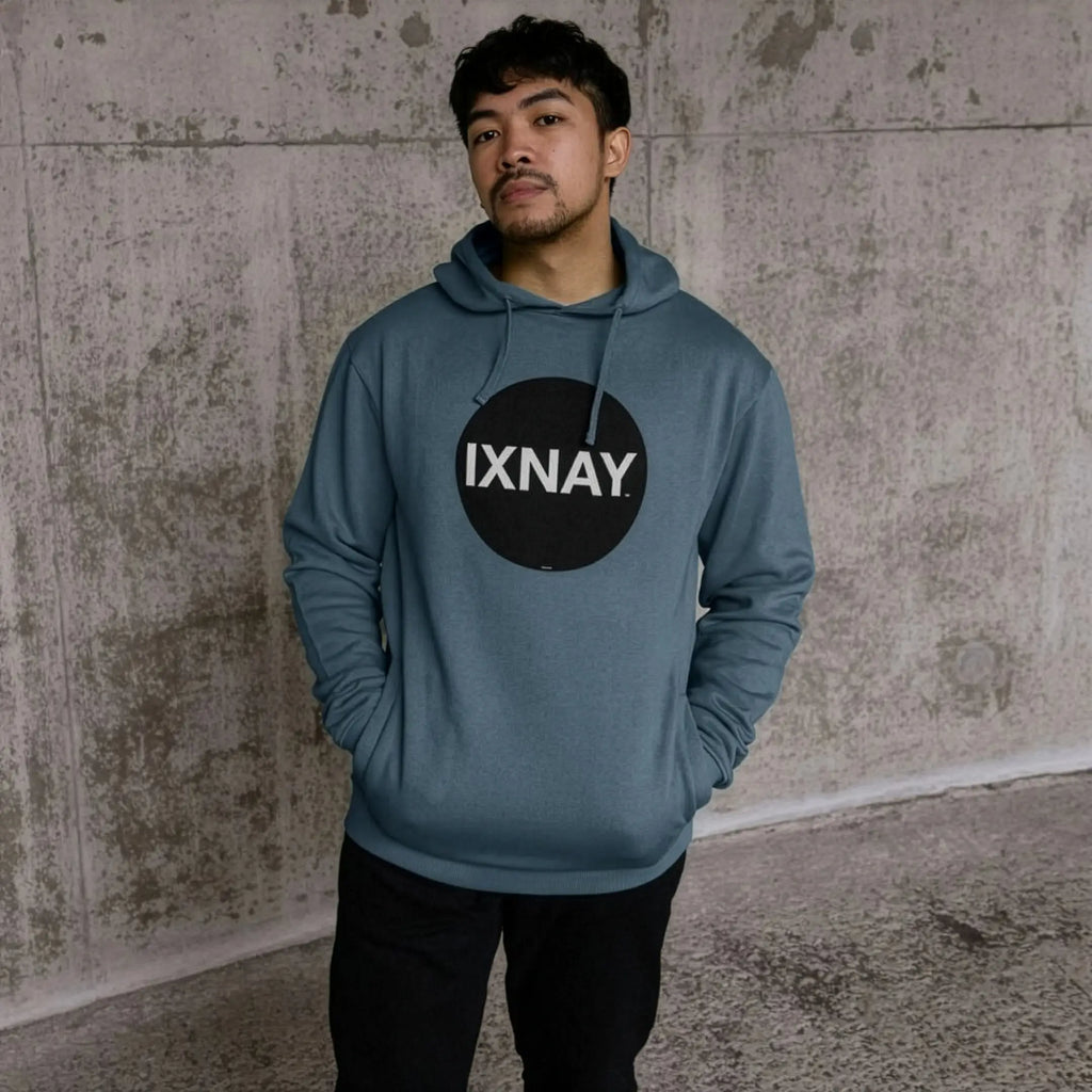 Man in urban concrete structure wearing mens stone blue organic cotton pullover hoodie with original black round IXNAY logo.