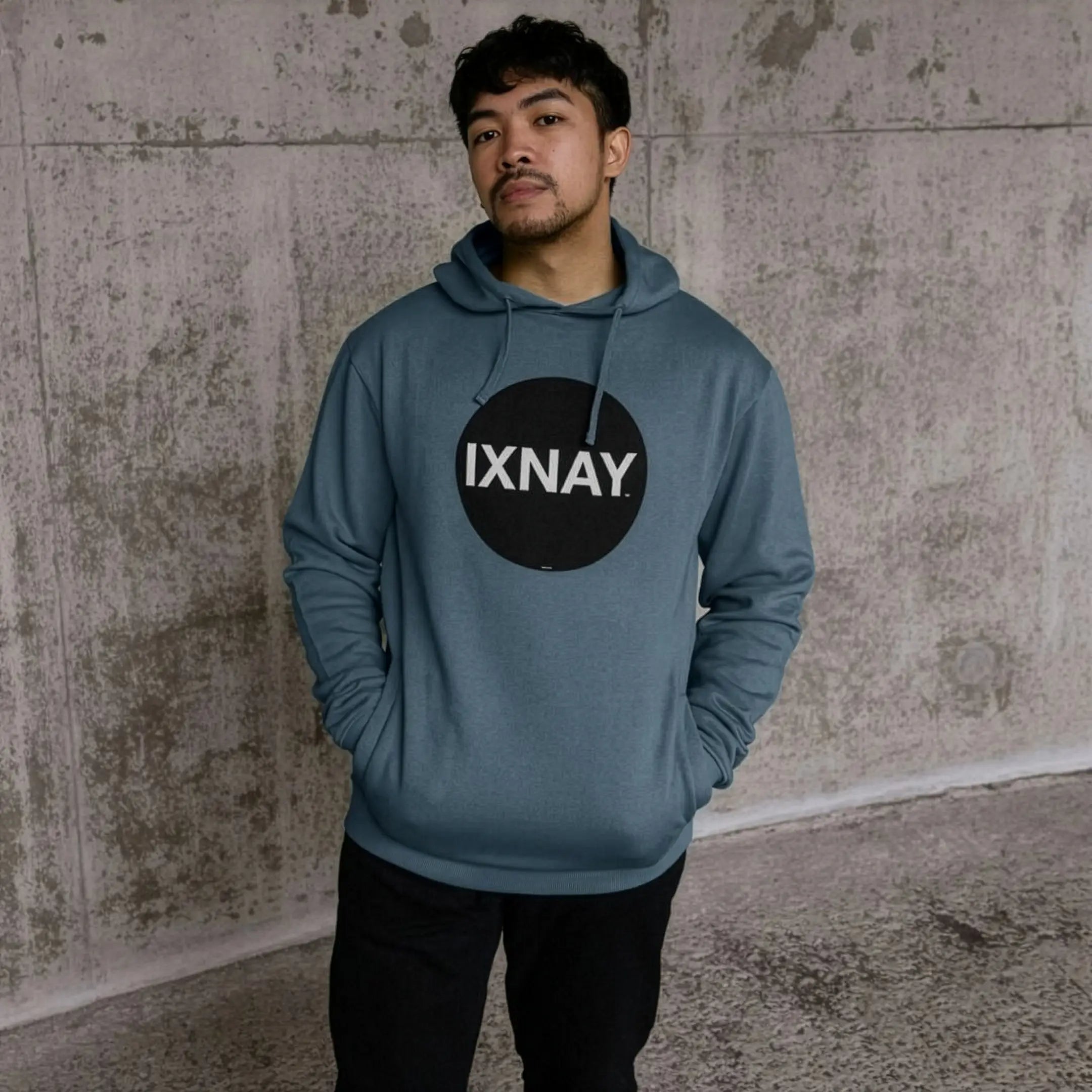 Man in urban concrete structure wearing mens stone blue organic cotton pullover hoodie with original black round IXNAY logo.