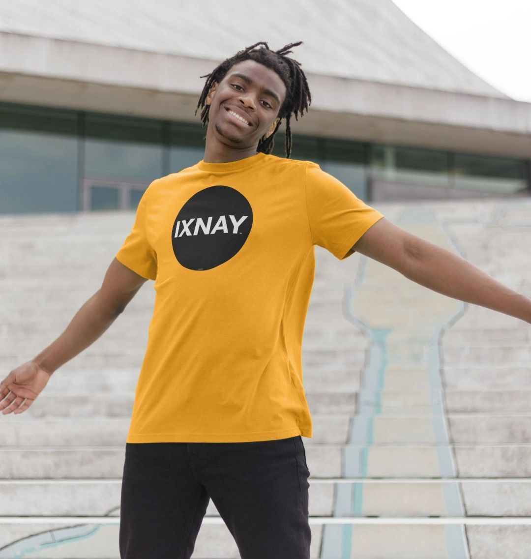 Black man standing on the wide concrete steps of a modern public building wearing a mustard yellow Printed Men's T-Shirt with Original Black Round IXNAY Logo with the phrase REJECT REJECTS REJECTOR on back  to represent the artist and musician IXNAY