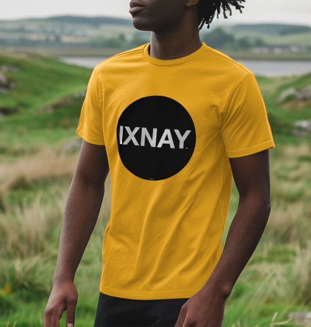 A black man standing among grass with a bay in the distance wearing a mustard yellow Printed Men's T-Shirt with Original Black Round IXNAY Logo with the phrase REJECT REJECTS REJECTOR on back  to represent the artist and musician IXNAY