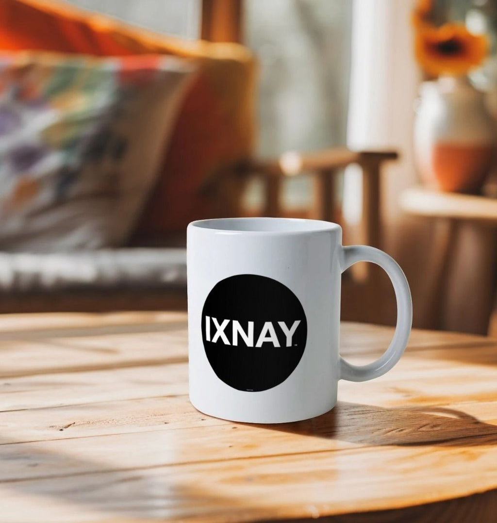 white ceramic mug emblazoned with the black logo with white lettering for the artist and musician IXNAY on a wooden table in a room bathed in warm light with a sofa and table in the background with typical house furnishings in white, orange and beige