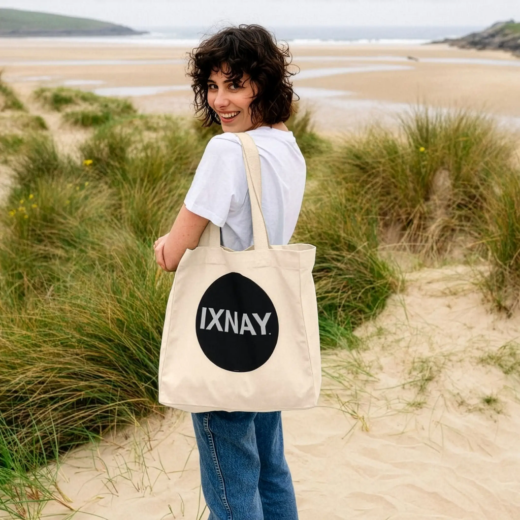Woman at beach holding IXNAY Original logo 100 percent organic natural cotton tote bag over her shoulder.