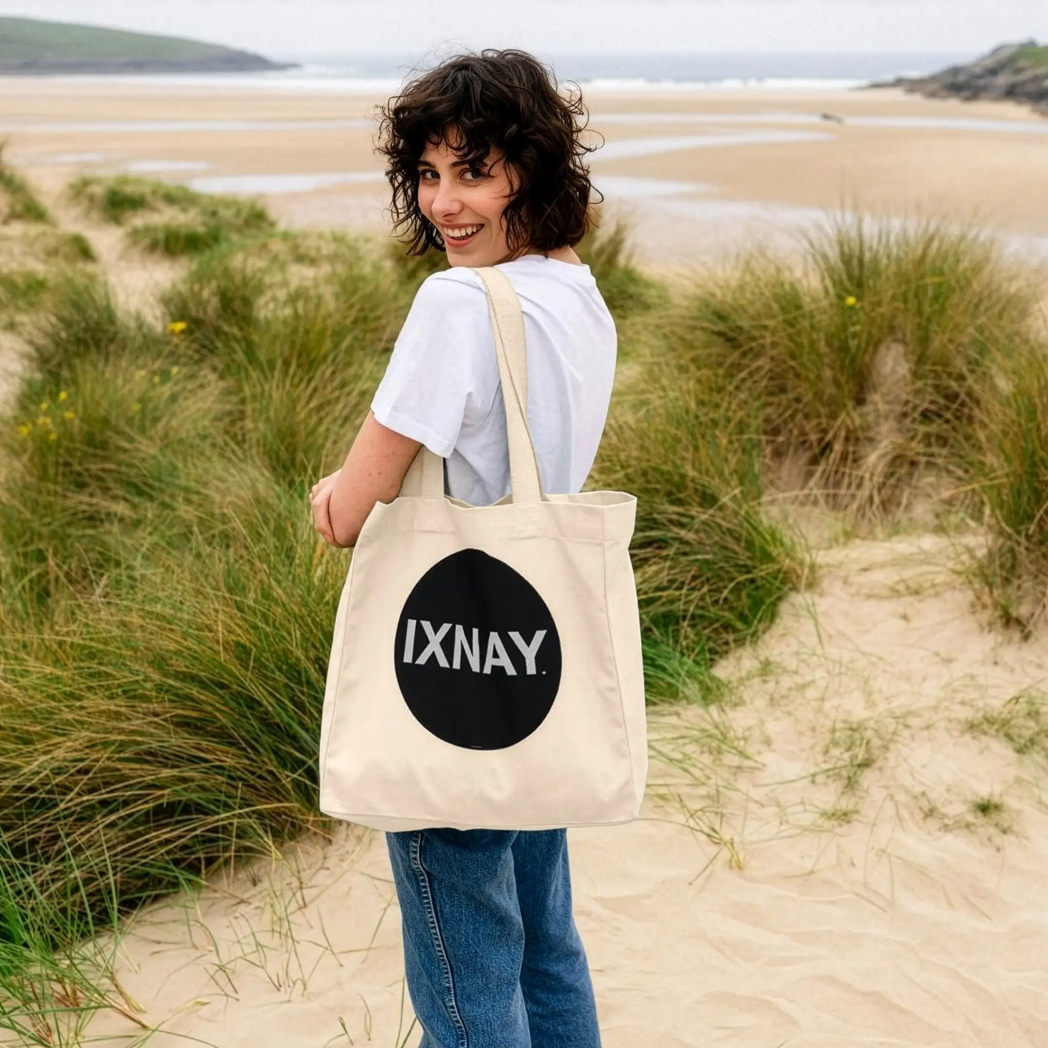 Woman at beach holding IXNAY Original logo 100 percent organic natural cotton tote bag over her shoulder.