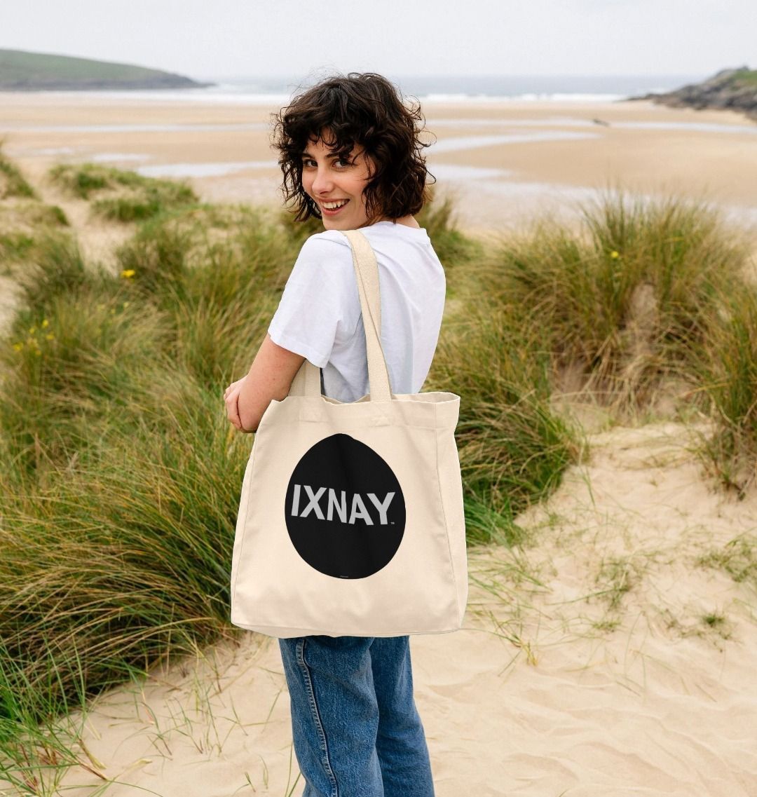 woman at beach over the shoulder view of IXNAY original round logo tote bag natural 100% organic cotton canvas