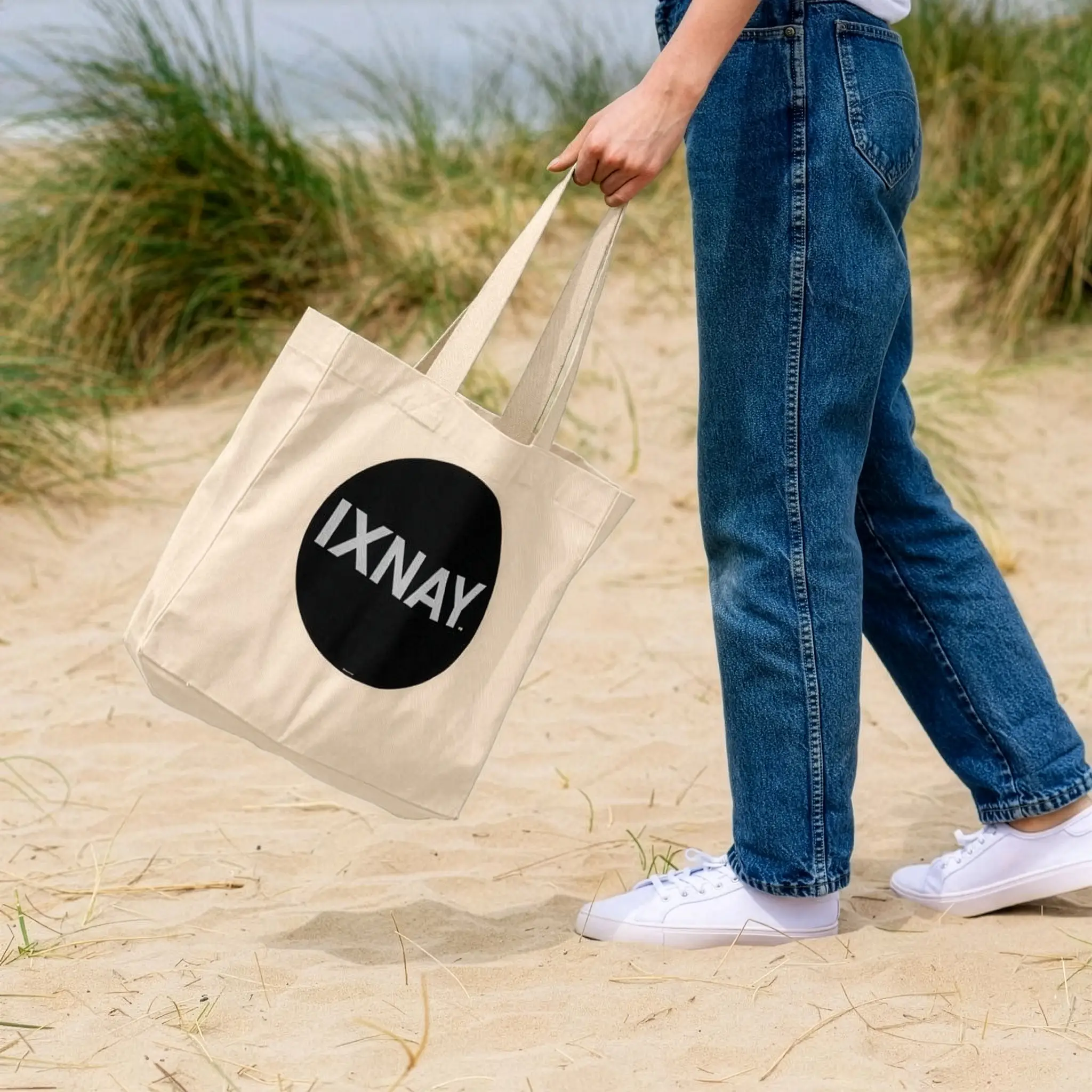 Woman at beach swinging an IXNAY Original logo 100 percent organic natural cotton tote bag by the reinforced strap handle.