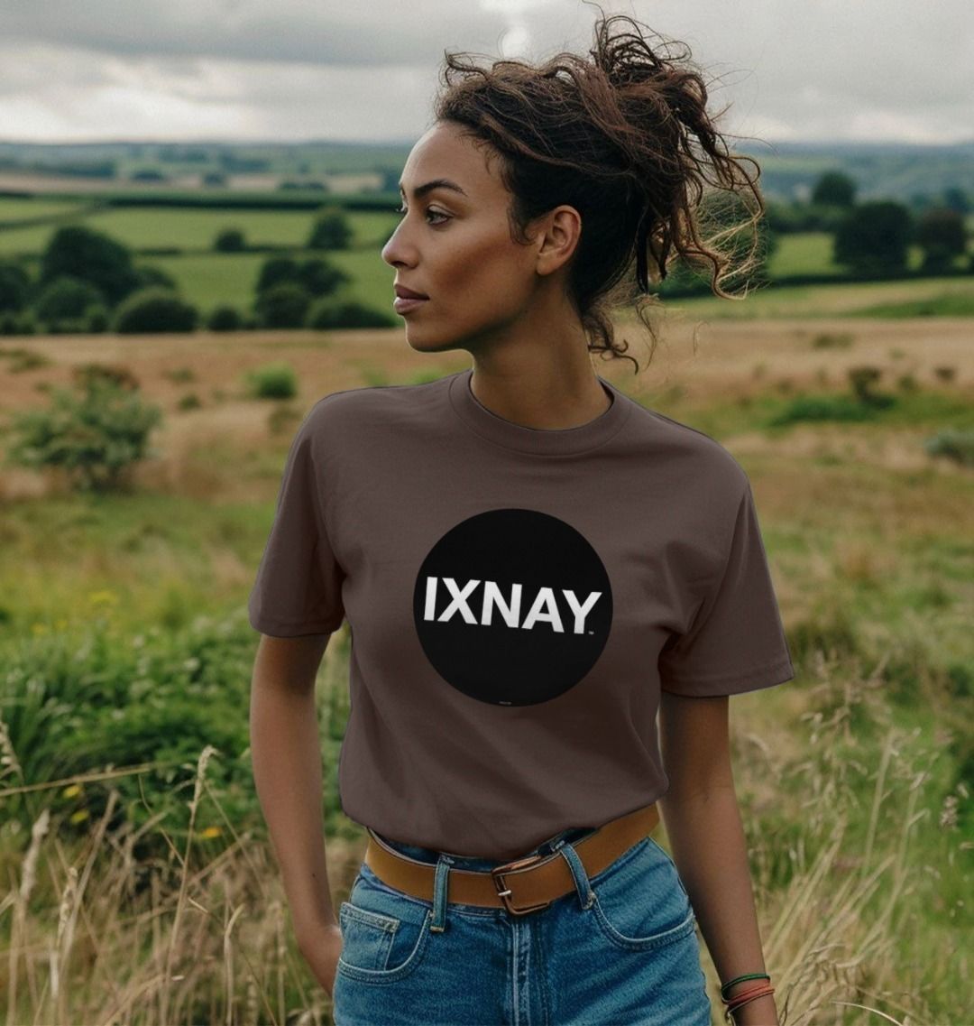 Woman striking a dramatic profile pose and standing in meadow with lush fields in the distance wearing a chocolate brown Printed Women's T-Shirt with Original Black Circular IXNAY Logo