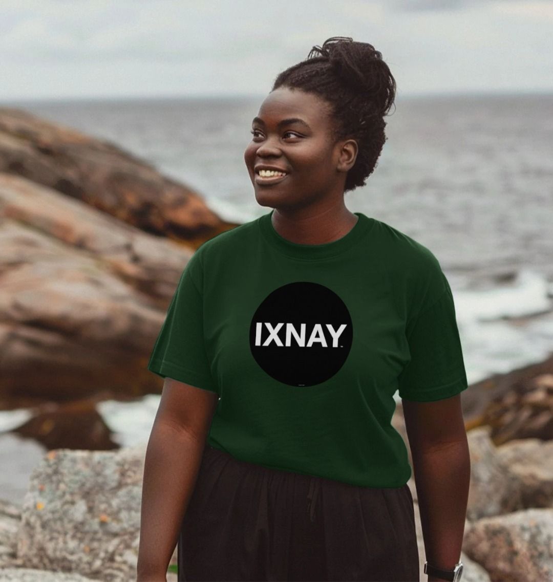 A Black woman smiling while standing on a rocky ocean beach wearing an evergreen colored Printed Women's T-Shirt with Original Black Circular IXNAY Logo