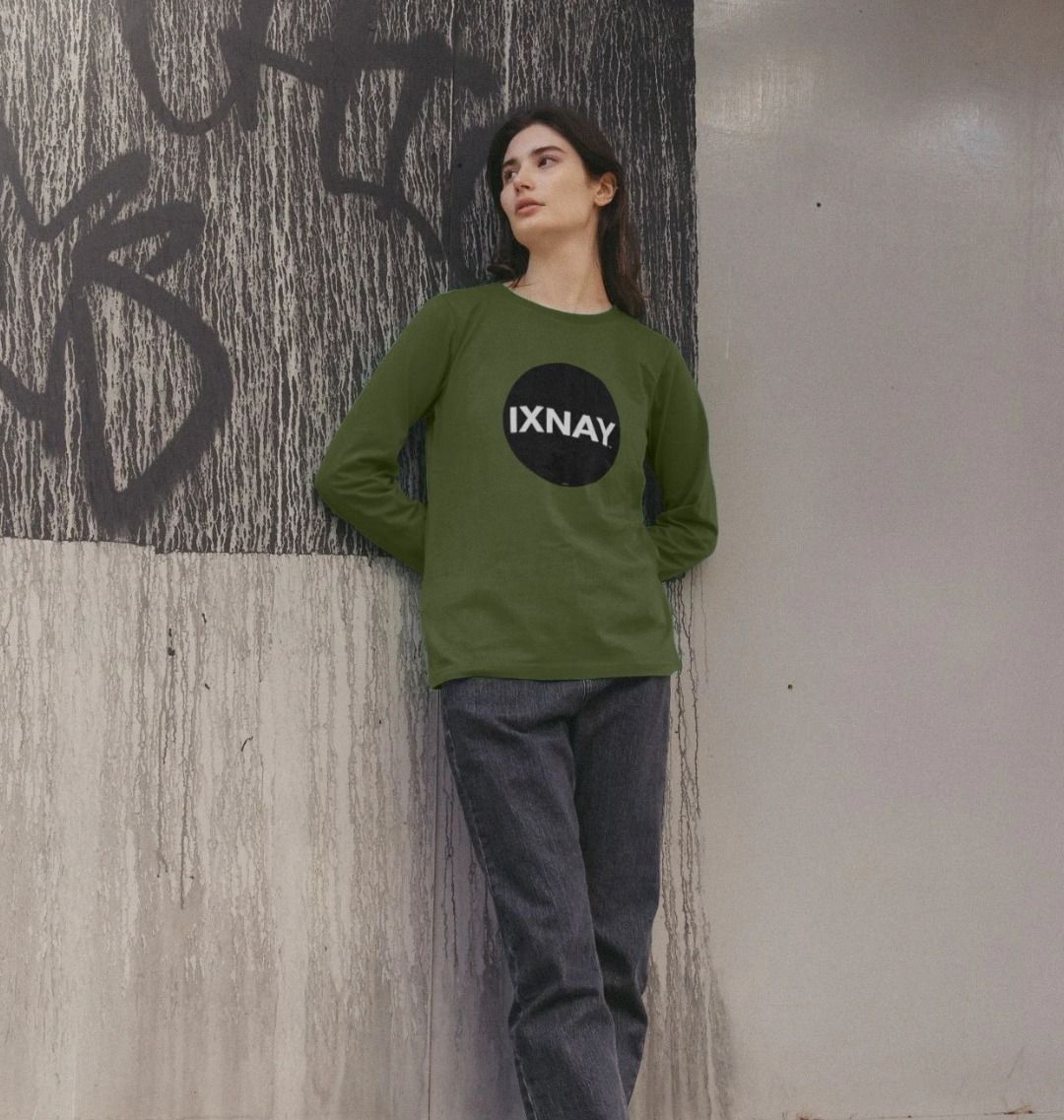 Woman in front of graffitied cement wall wearing khaki green Printed Women's Long-sleeved T-Shirt with Original Black Round IXNAY Logo from the artist and musician IXNAY