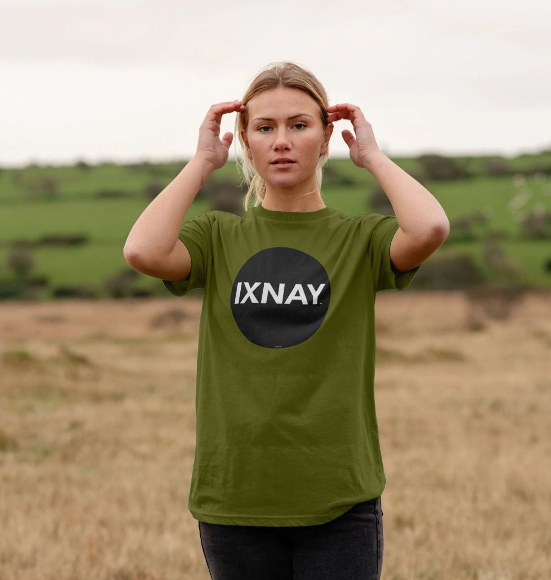 Blond woman standing in a field wearing an moss green Printed Men's T-Shirt with Original Black Round IXNAY Logo with the phrase REJECT REJECTS REJECTOR on back  to represent the artist and musician IXNAY