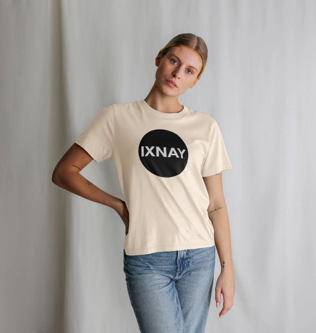 Woman striking a pose and standing in front of a draped white cloth background wearing a light oat beige Printed Women's T-Shirt with Original Black Circular IXNAY Logo