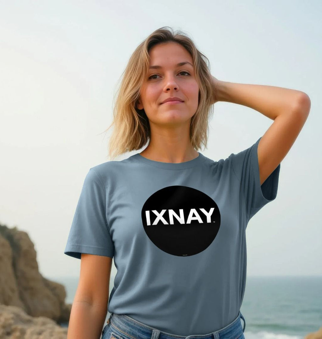 Woman smiling and standing on a rocky beach wearing stone blue Printed Women's T-Shirt with Original Black Circular IXNAY Logo