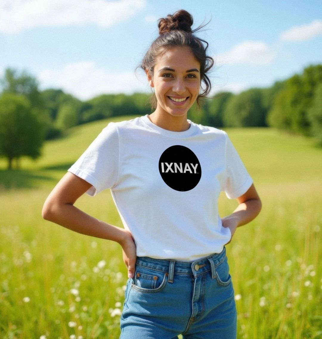 Woman smiling and standing in meadow with a forest tree line in the background wearing a white Printed Women's T-Shirt with Original Black Circular IXNAY Logo