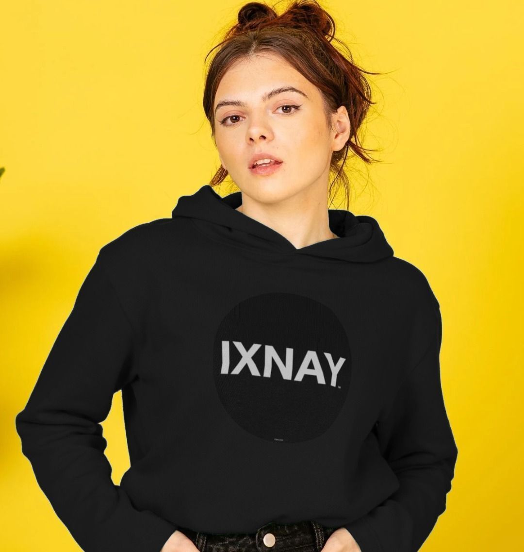 woman in front of bright yellow background wearing womens black organic pullover hoodie with original black round IXNAY logo