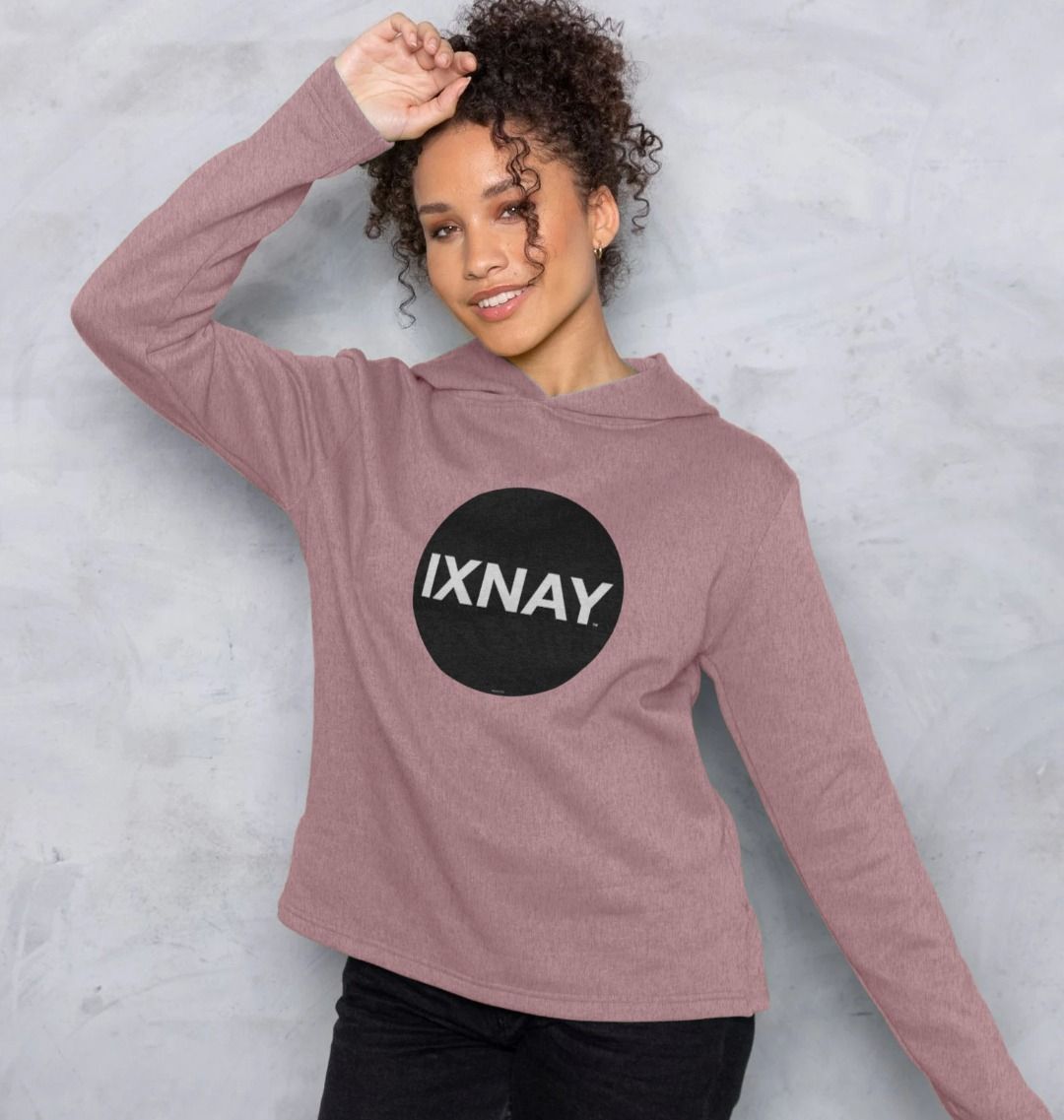 woman in front of cement wall wearing womens sunset pink organic remill pullover hoodie with original black round IXNAY logo